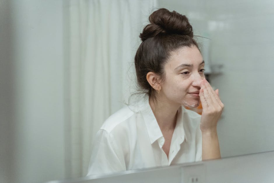 Woman applying face cream in bathroom, focusing on skincare routine with natural reflection in mirror.