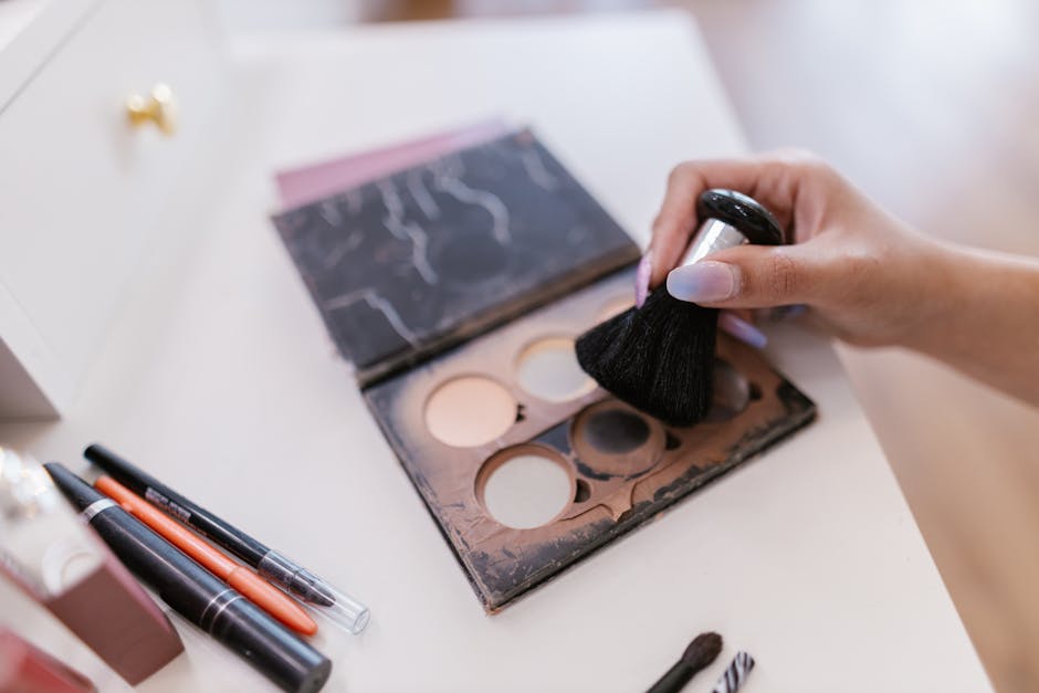 A hand holding a makeup brush over an open palette with brushes nearby on a table.