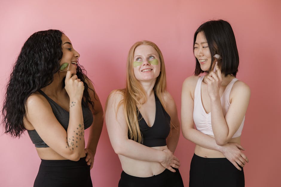 Three young women using jade rollers and eye patches on a pink background.