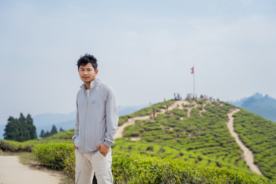 Asian man in casual attire posing on a scenic hill with lush greenery under a clear sky.