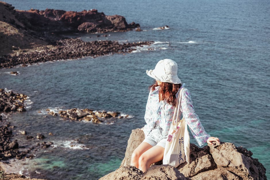 Young woman in summer attire sitting on rocks, overlooking a coastal landscape.