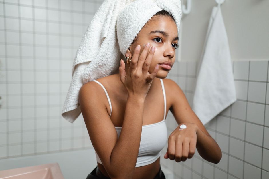 A young woman in a bathroom applying skincare cream with a towel on her head, focusing on self-care.