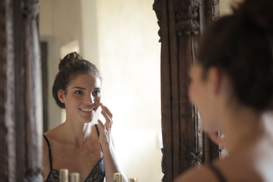 Young woman applying skincare and smiling at her reflection in a rustic mirror.