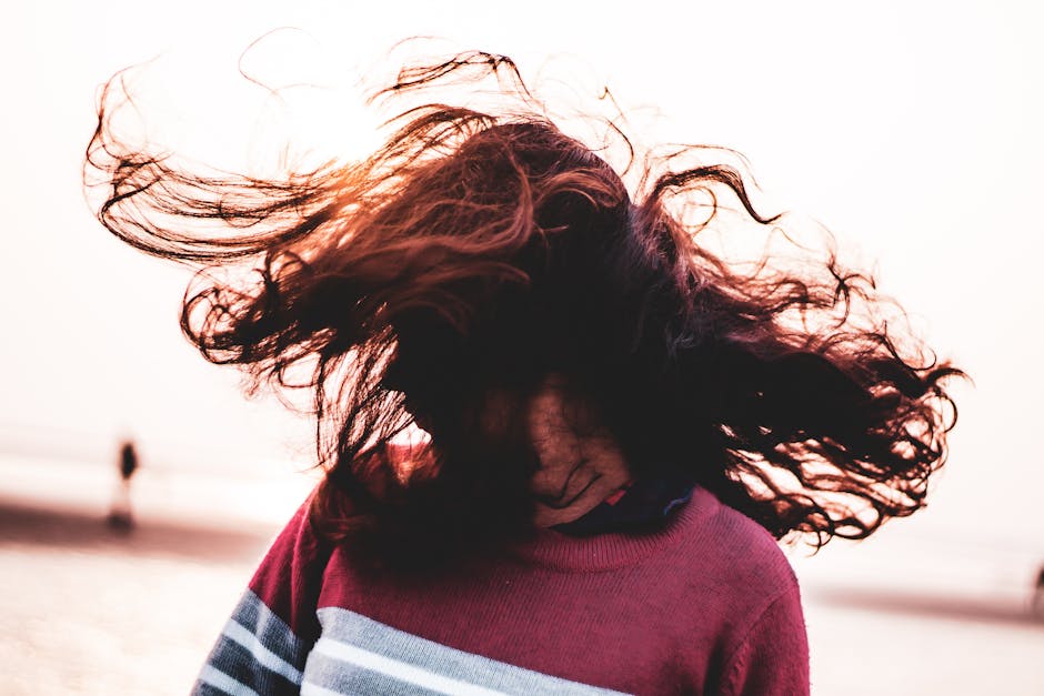 Anonymous female in sweater shaking head with long dark hair while standing on embankment near sea against blurred background in coastal area