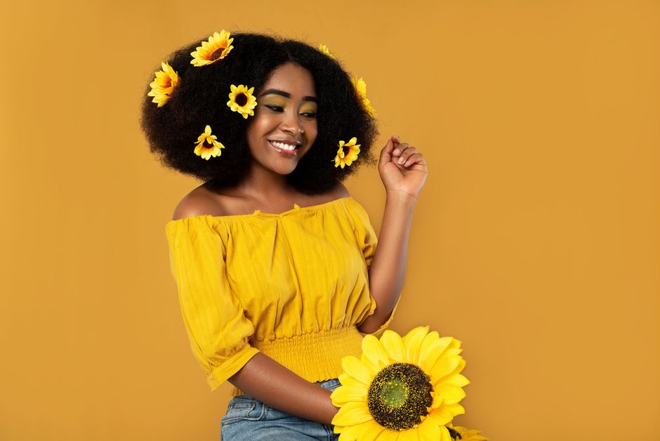 Smiling woman with sunflowers in hair against a vibrant yellow backdrop.