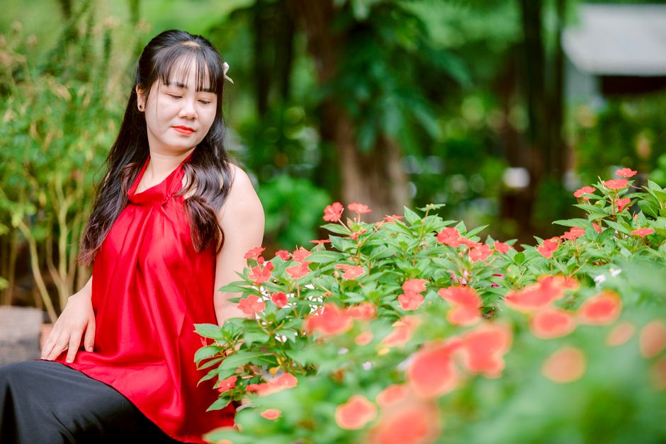 A woman in a red dress sitting serenely among lush green foliage and vibrant flowers.