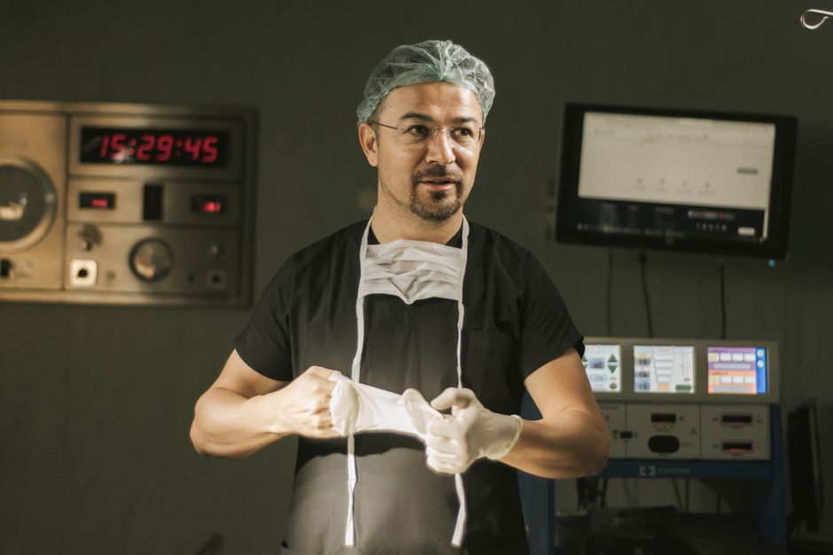 A male surgeon in scrubs and mask prepares for surgery in a hospital operating room.