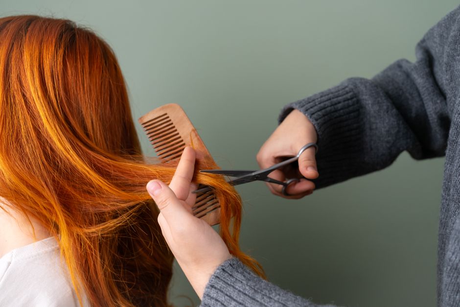 Close-up of red hair being trimmed with scissors and comb on green background.